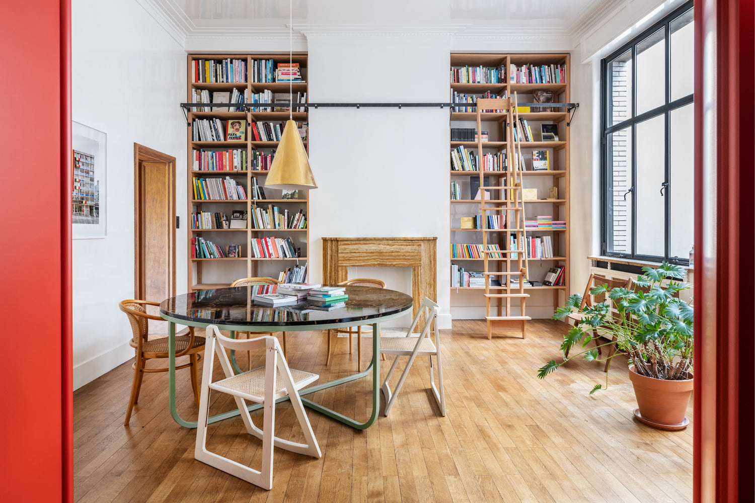 Library and meeting room at the Luster office in Antwerp. A round table with several chairs, a large window and two large bookcases in the back. Left and right red folding door panels. A potted monstera plant on the floor.